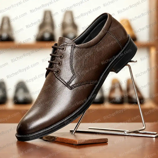 A brown leather lace-up dress shoe displayed on a metal stand against a blurred background of shelves with other shoes. The shoe has a pebbled leather texture, a rounded toe, and dark laces.